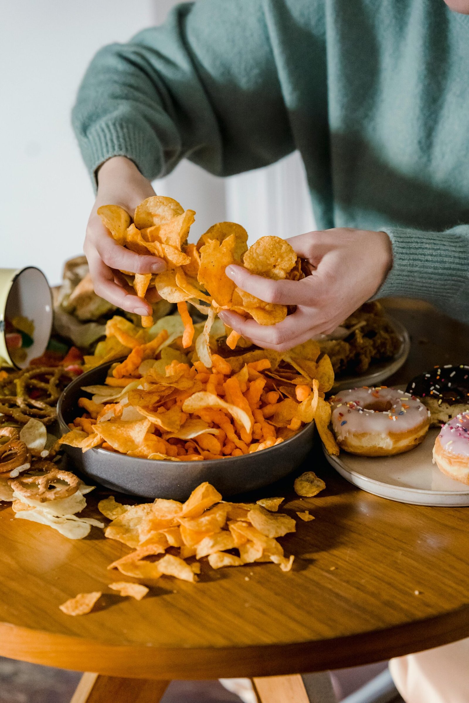 Unrecognizable female with tasty unhealthy crisps in hands sitting at table with various junk food in light kitchen at home